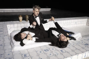 Drei Darsteller*innen in Anzügen sind auf einer Matratze in einem gefliesten Becken