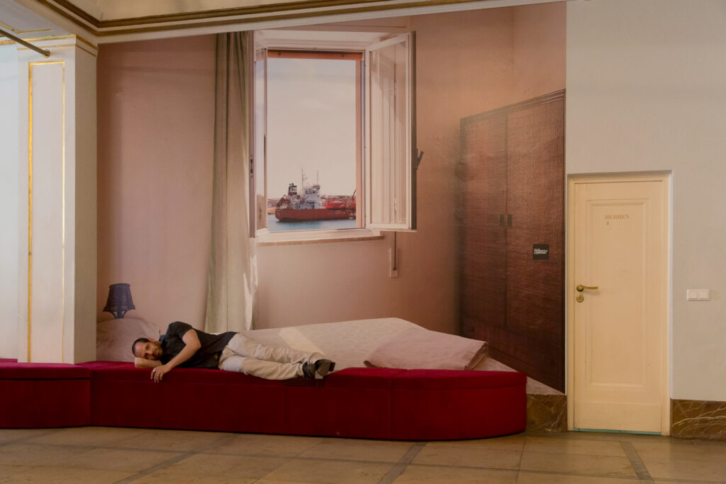Der Schauspieler Yousef Sweid im Maxim Gorki Theater. Im Hintergrund die Fotografie von Sven Johne: „Traumhotels (Lampedusa)“, Fotoserie 2012. Foto (c) Judith Buss.